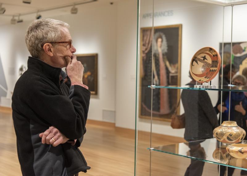 A white man wearing glasses stands in an art gallery looking at pottery in a glass case