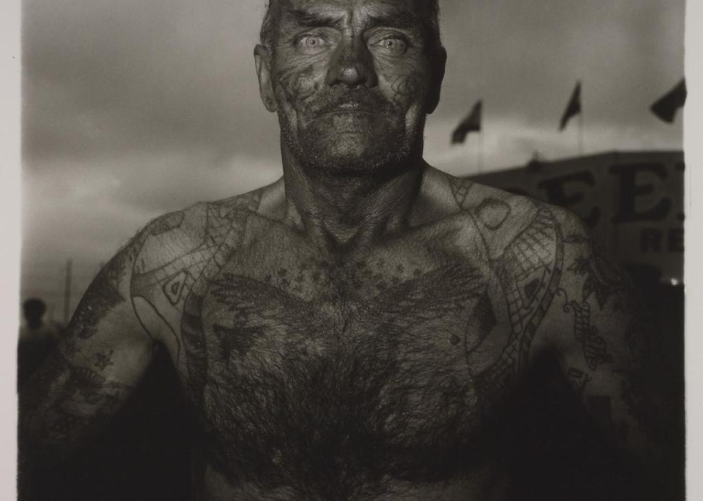 A black-and-white photo of a shirtless, balding white man with tattoos on his face and chest staring out toward the viewer