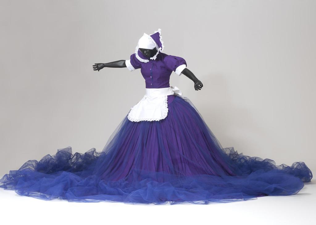 An African American female mannequin stands with outstretched arms wearing a purple and blue dress with a large skirt and a white maid’s apron