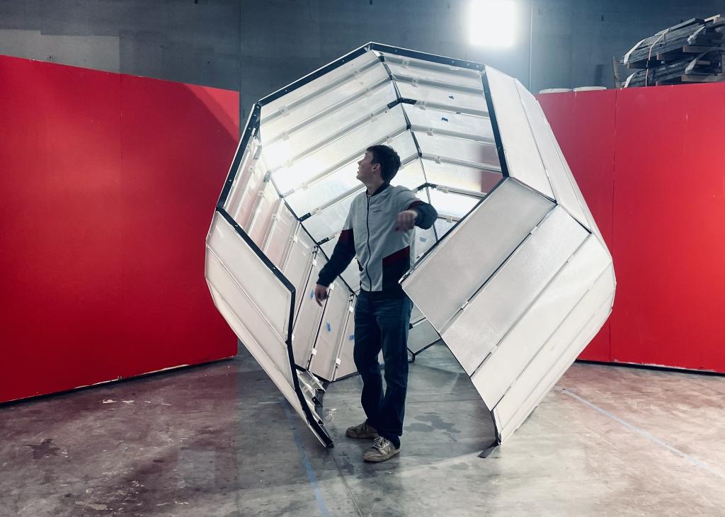 An adult stands inside a six-sided sculpture with an opening that rests on the ground of an industrial space