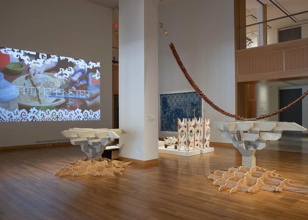 A gallery with wood floors and white walls with concrete sculptures and a large video projected on one wall