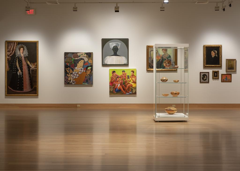 Art gallery with wood floors and white walls displaying bright paintings of women through the centuries
