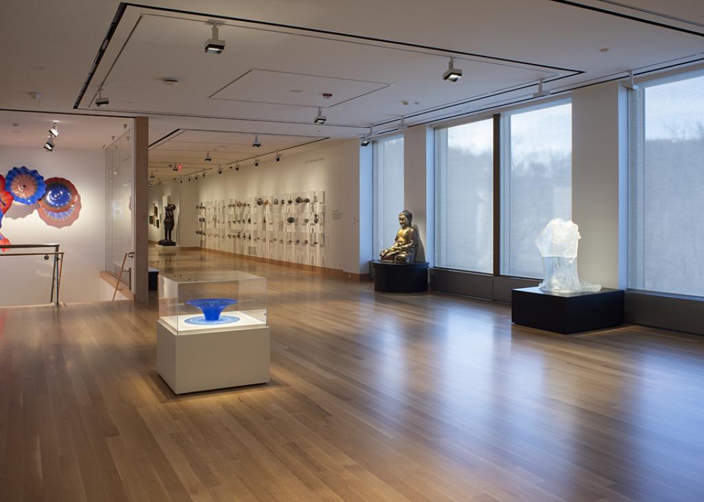 Photograph of a dimly lit art gallery with wooden floors and large windows; multiple sculptures line the walls 