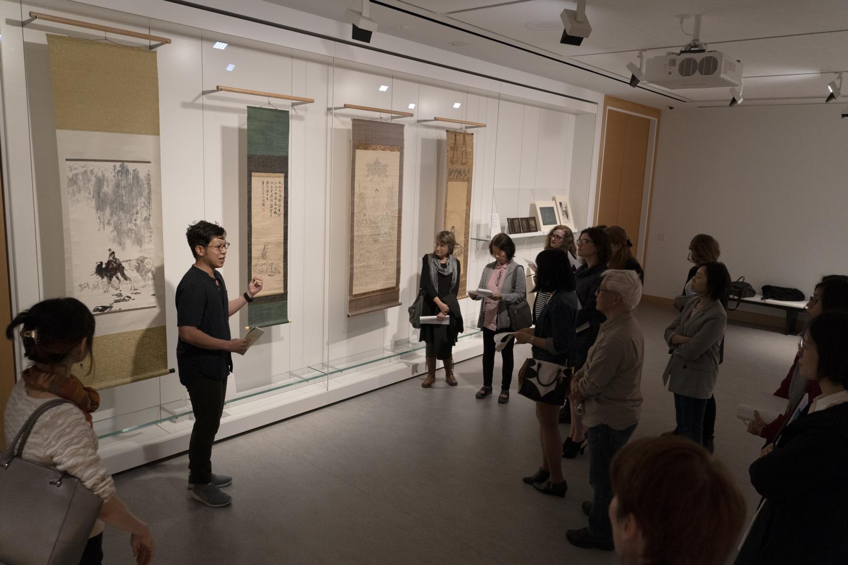 Adults stand in a dark art gallery around a man gesturing toward large scrolls hanging on the walls behind him