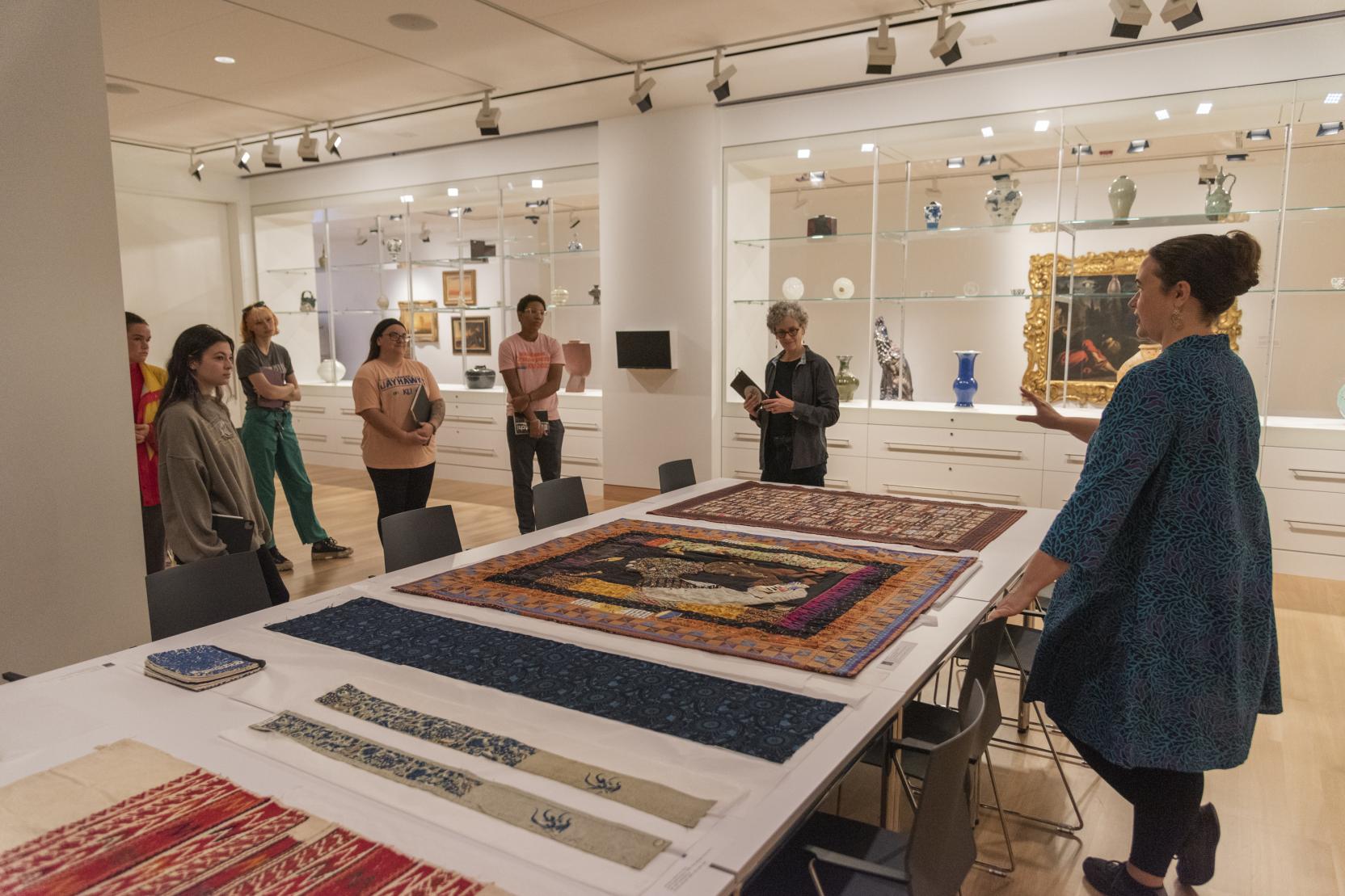 Adults stand in a room with glass walls and look toward a woman gesturing to artworks lying on a table