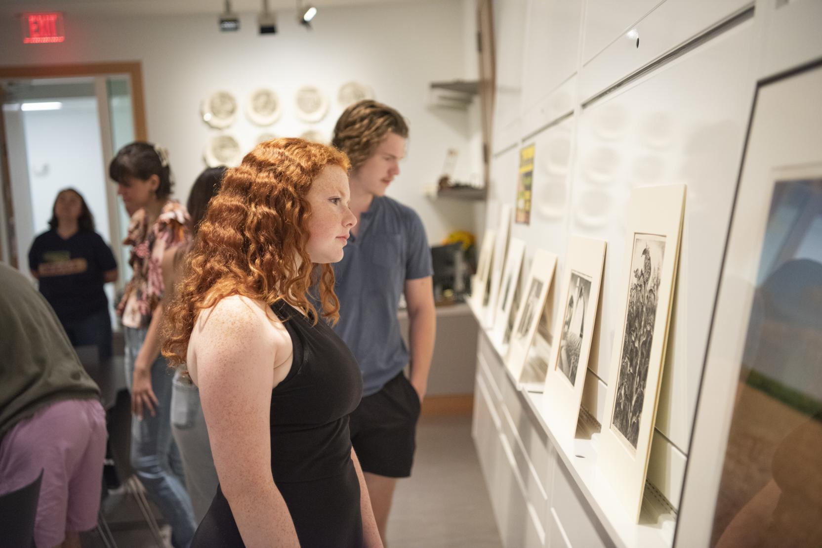 A woman with red curly hair stands in a gallery and looks toward art on the wall