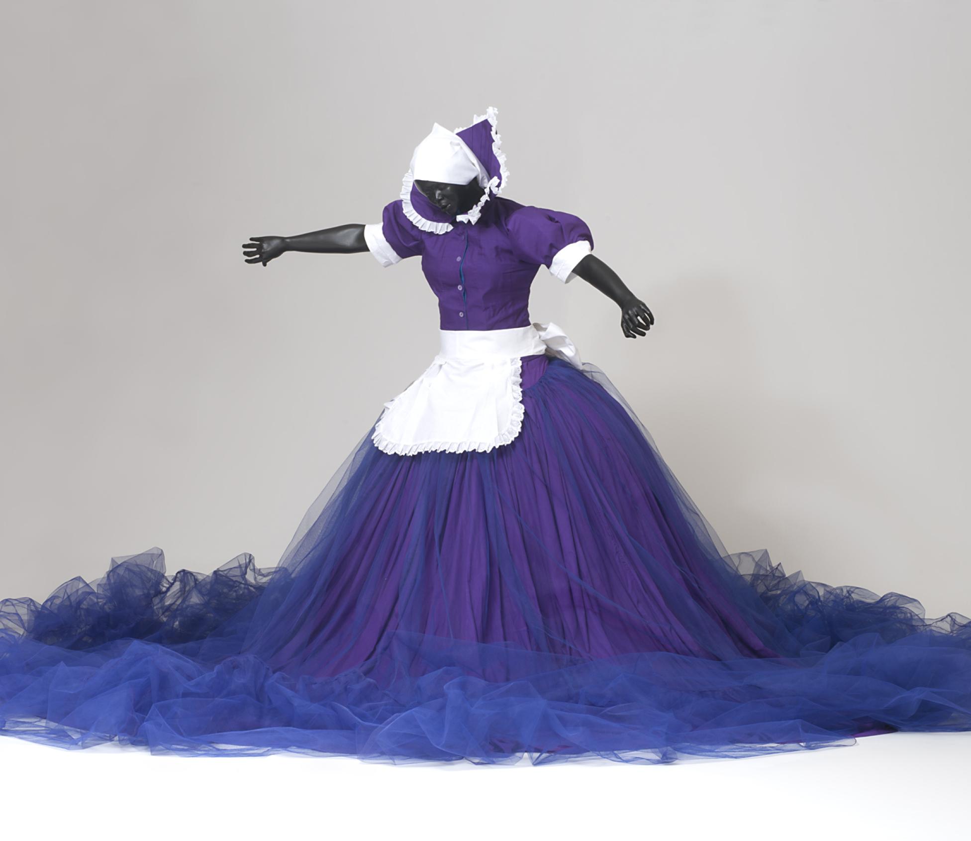 An African American female mannequin stands with outstretched arms wearing a purple and blue dress with a large skirt and a white maid’s apron 