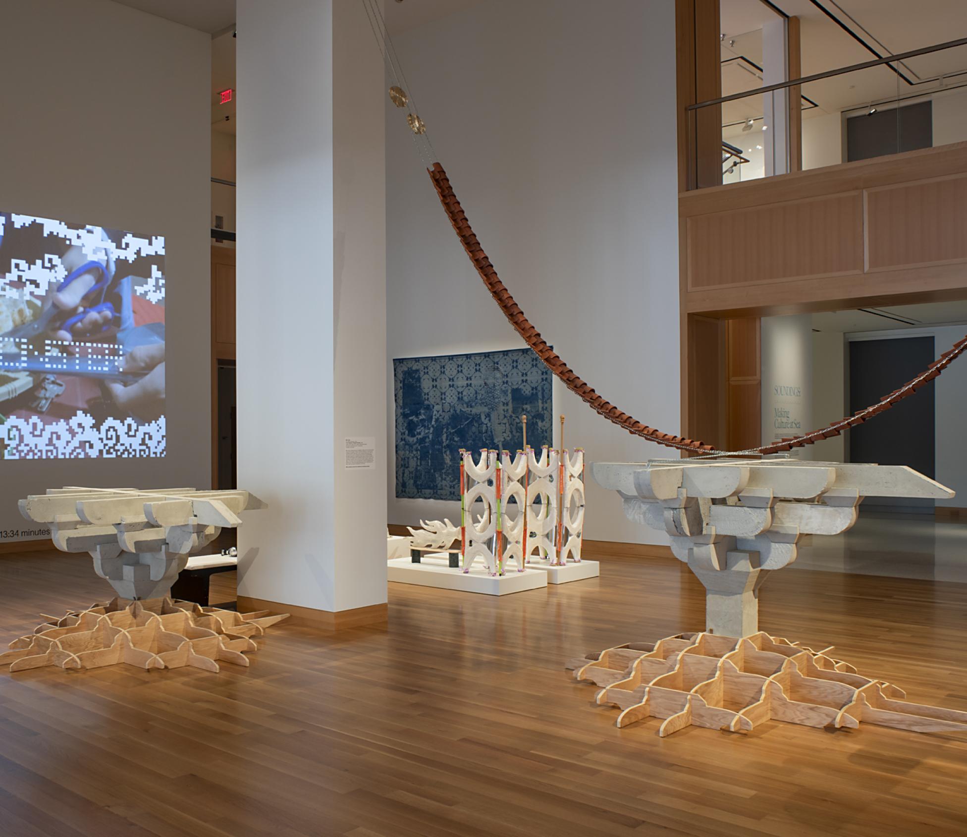 A gallery with wood floors and white walls with concrete sculptures and a large video projected on one wall