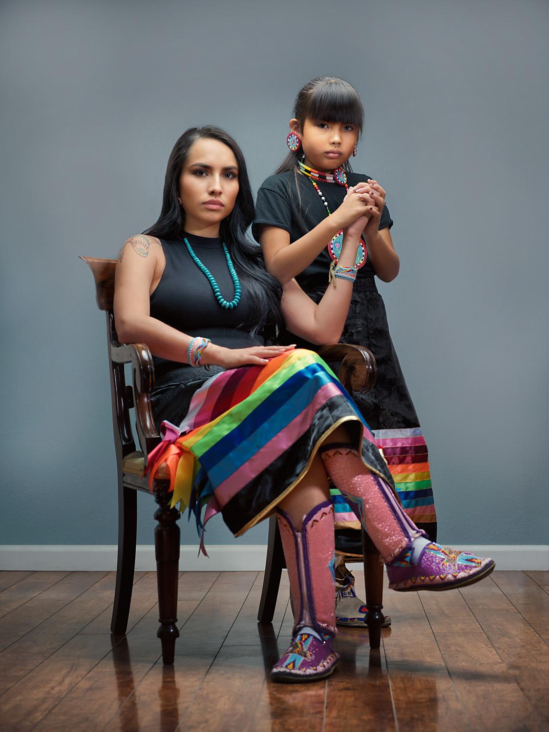 A seated Indigenous woman and standing girl hold hands in a formal pose staring at the camera; both wear black tops and colorful ribbon skirts and accessories