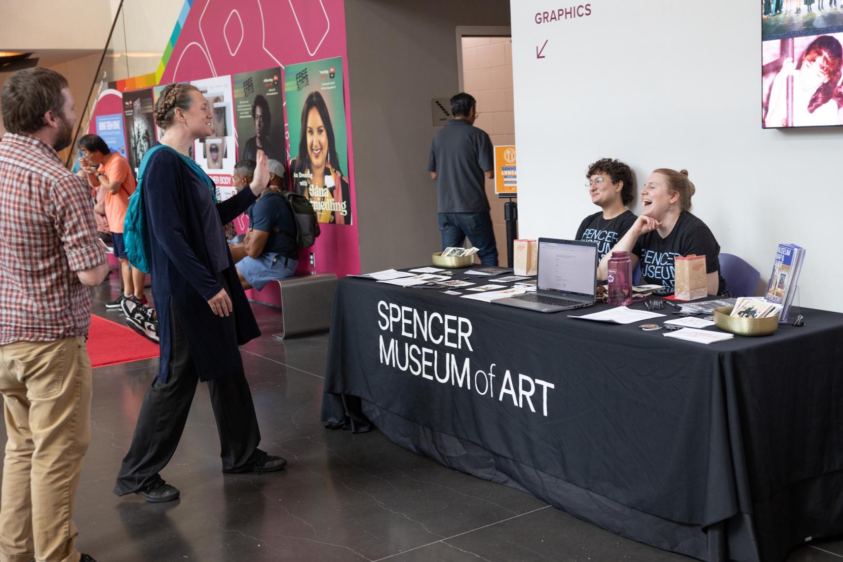 Two adults sitting at a table with a "Spencer Museum of Art" tablecloth talk with two other adults approaching the table