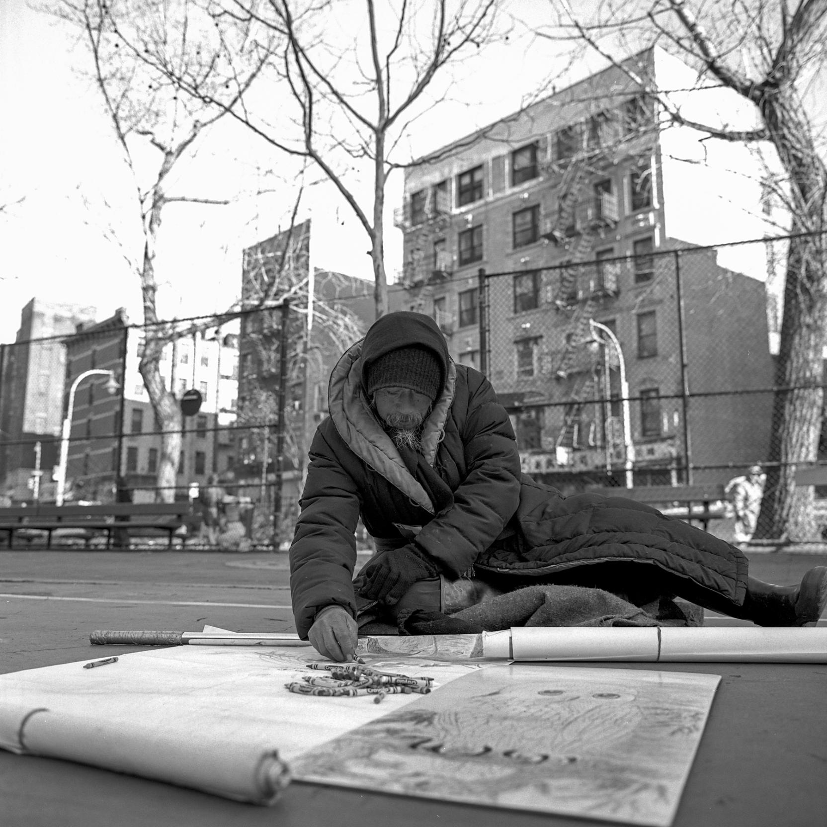 A man in a winter coat sits on the ground in a park and draws on a large piece of paper with crayons