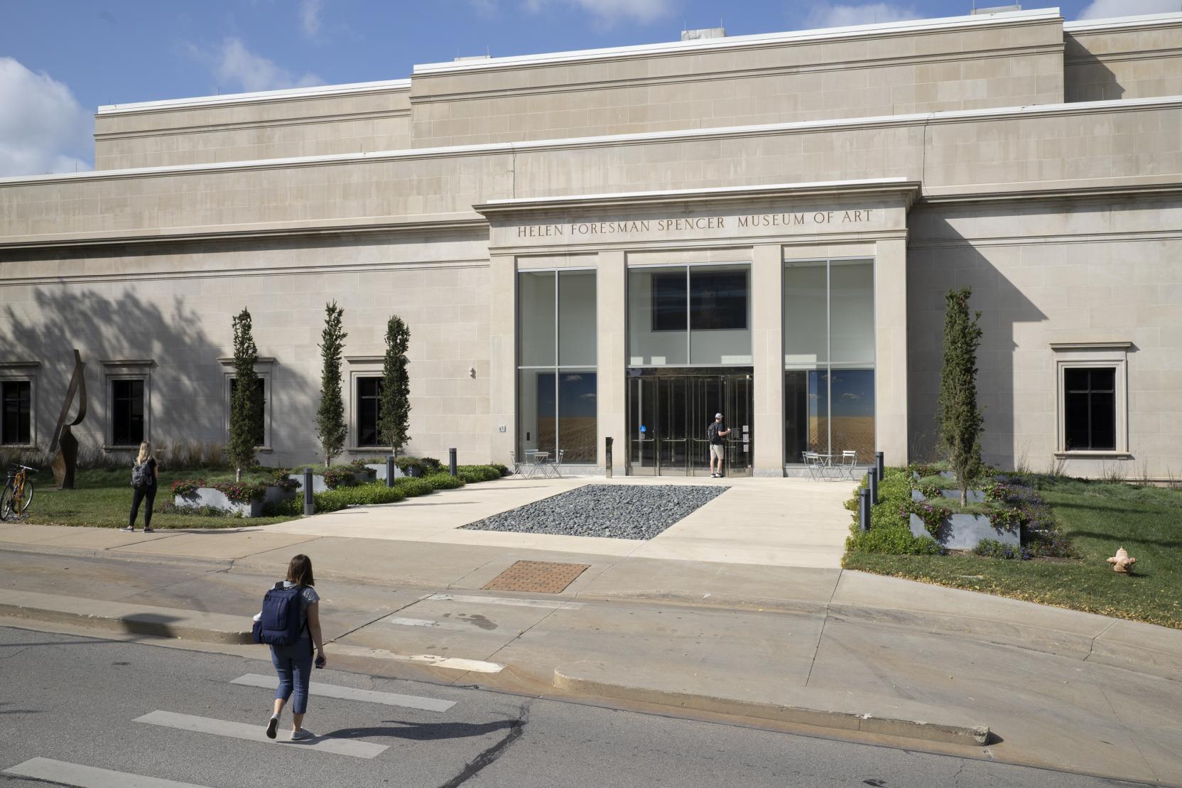 A woman wearing a backpack walks toward a large classic style building; “Helen Foresman Spencer Museum of Art” is displayed above the entrance