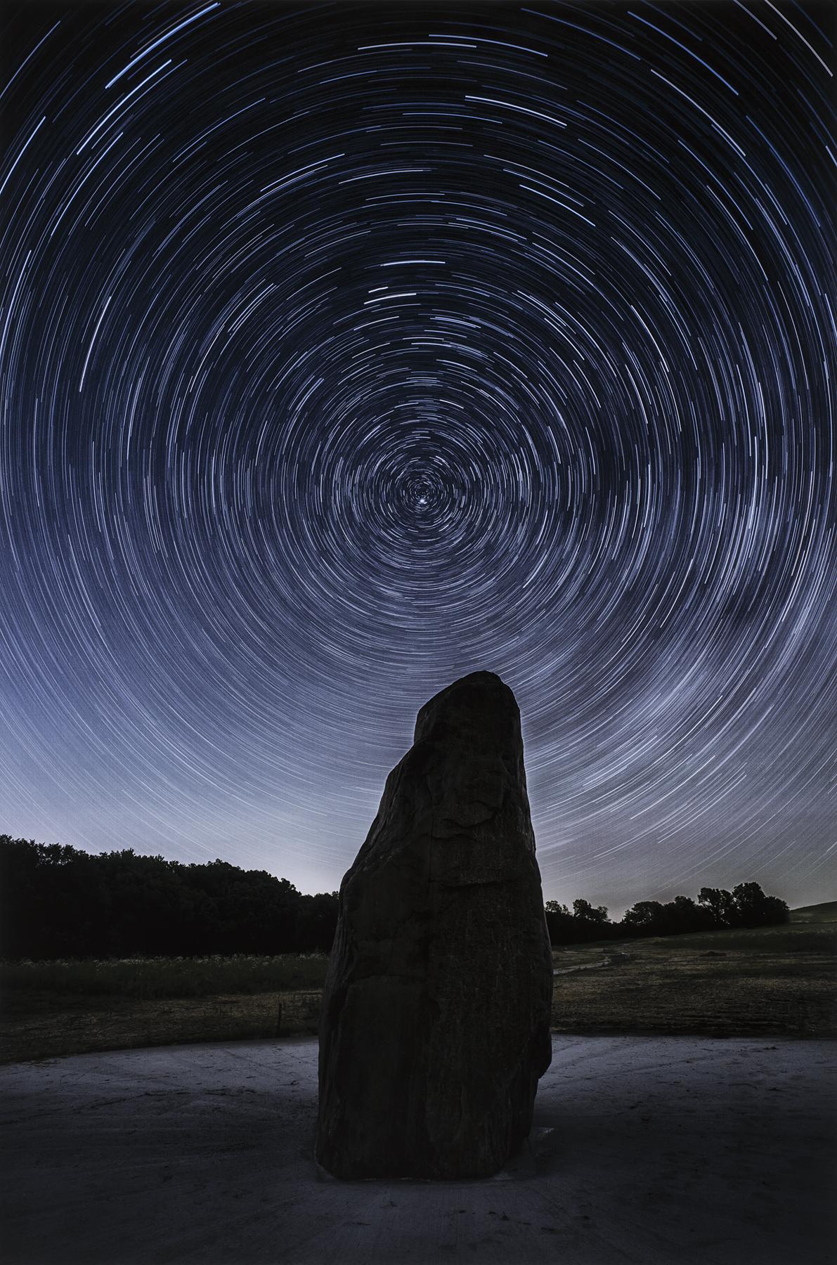 Stars swirl in a dark blue sky above a silhouette of a large rock in a long exposure photograph