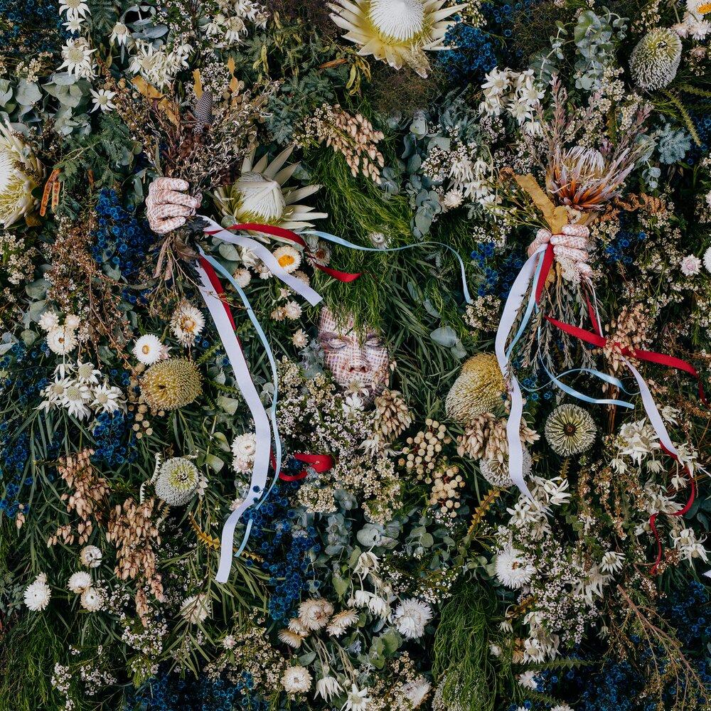amidst a dense wall of a variety of plants, a male face is barely visible; two hands also peak out holding bouquets died with red, white, and blue ribbons
