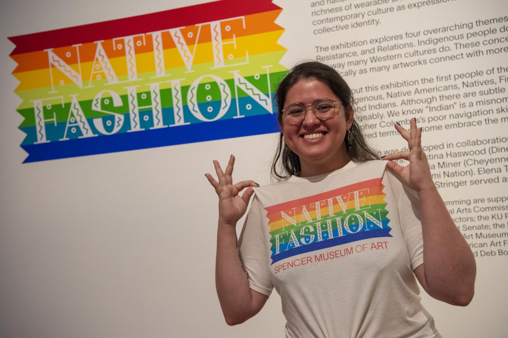 A young adult smiles at the camera while holding out their t-shirt that says "Native Fashion" in rainbow colors; the same design is on the wall behind them