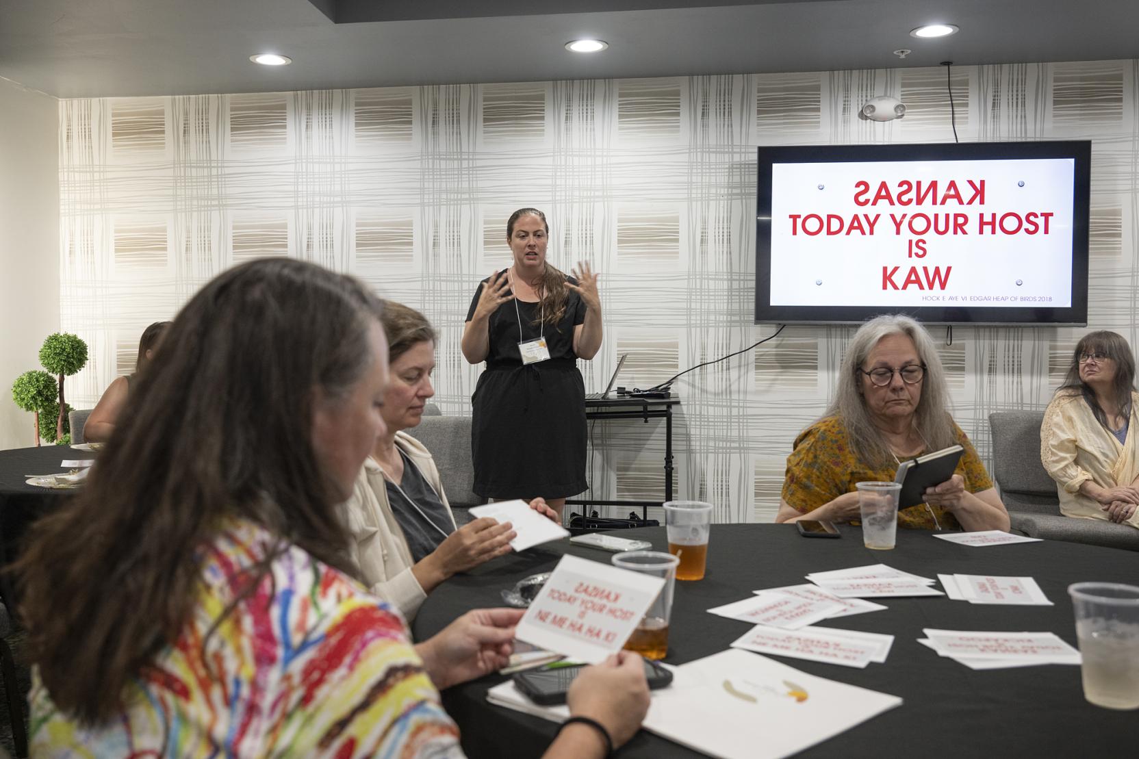 Several white women sit around a black table looking at notecards while another woman stands behind them