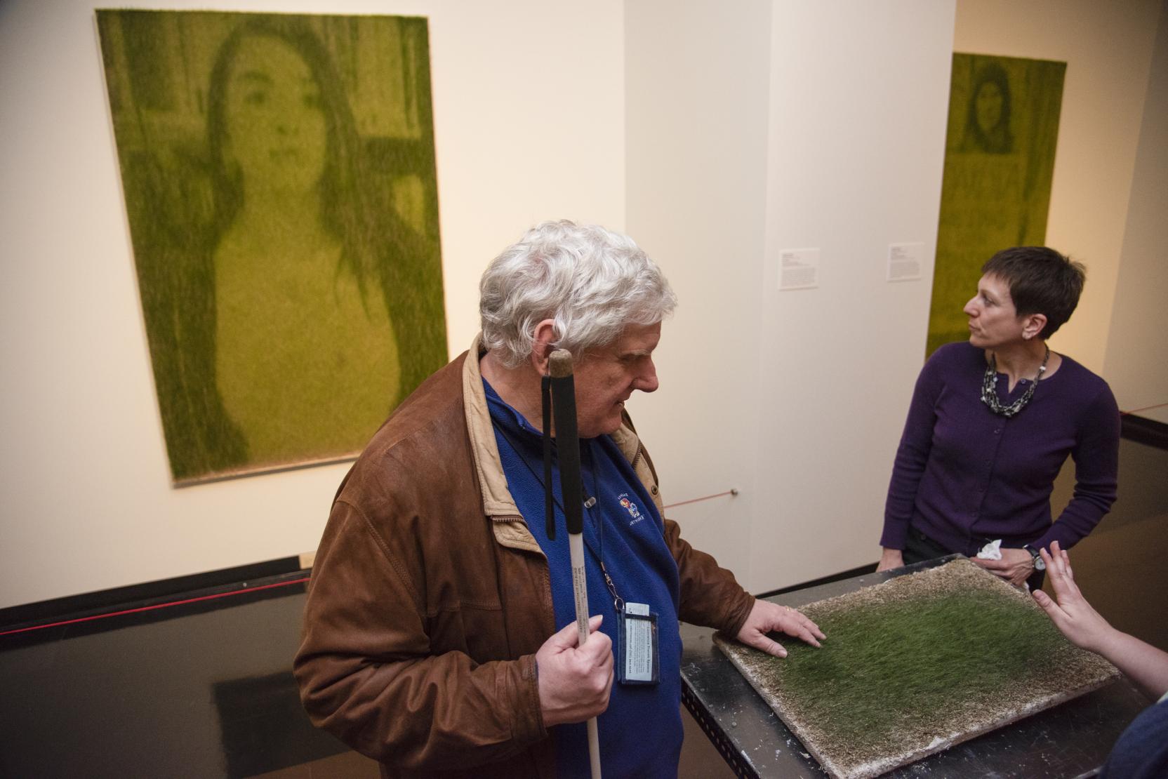 An elderly man holding a cane touches a painting on a table, a woman stands on the other side of the table