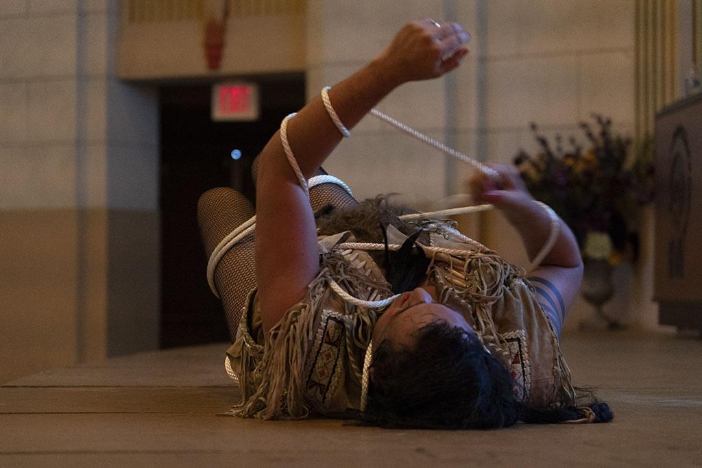 An Indigenous man lays on a stage holding a rope wrapped around his arms; the top of his head is toward the camera