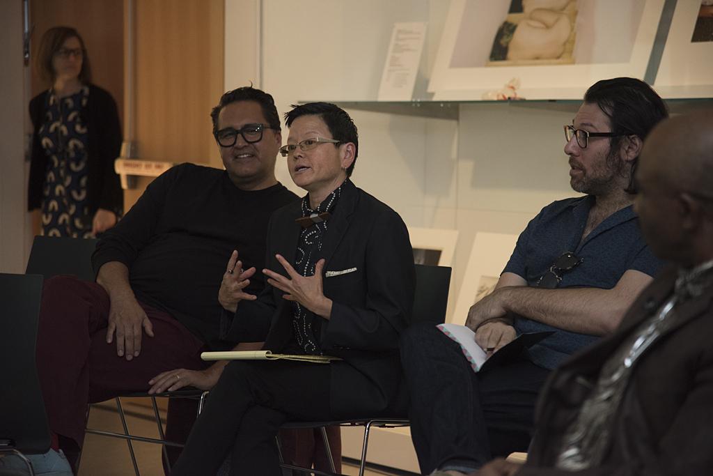 An Asian woman with close-cropped black hair sits between two men while talking with her hands