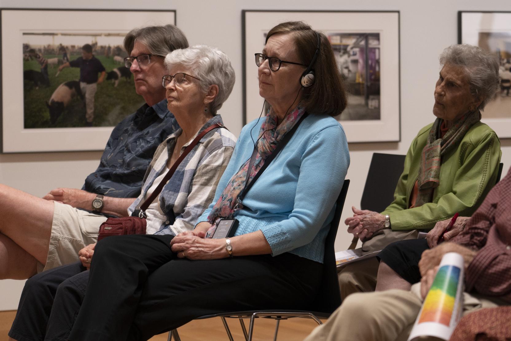 In a room full of seated people, an older woman wears over-the-ear headphones and looks forward at something out of frame