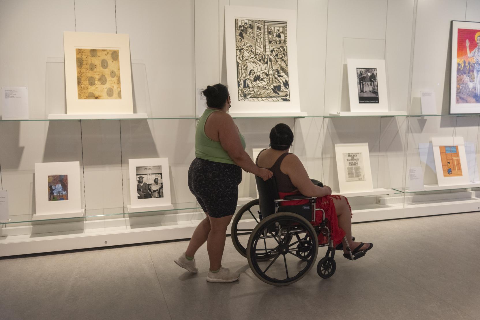 A woman stands behind a woman in a wheelchair; they both look toward a wall of art with their backs to the camera