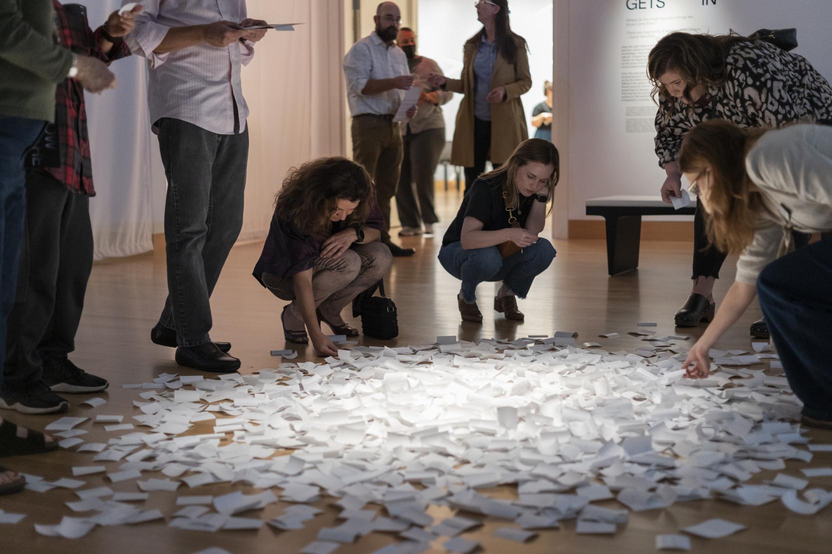 A group of adults hunch over a pile of small white papers spread across a gallery floor