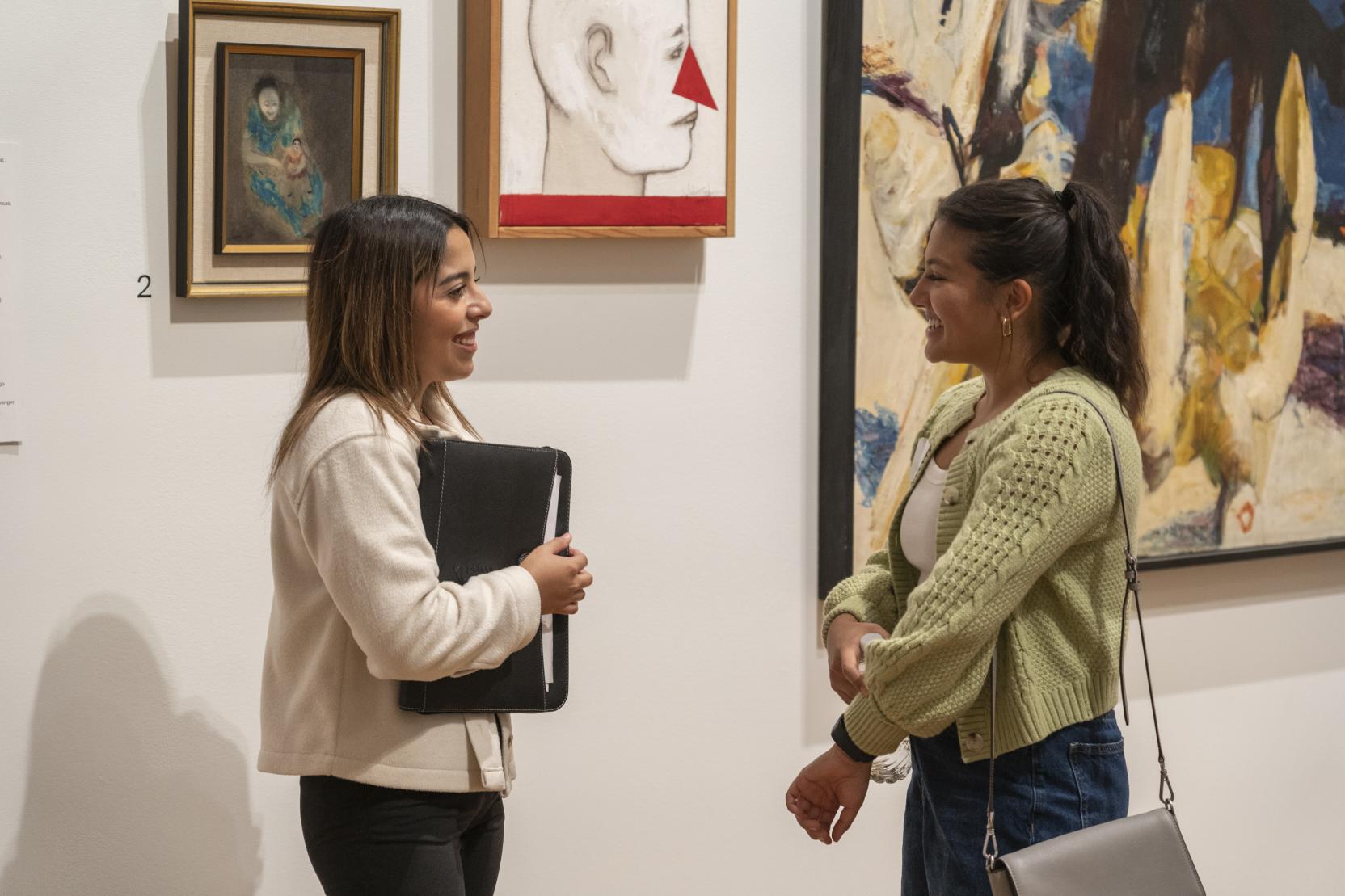 two young women, both with long brown hair and wearing sweaters, stand chatting in a gallery space