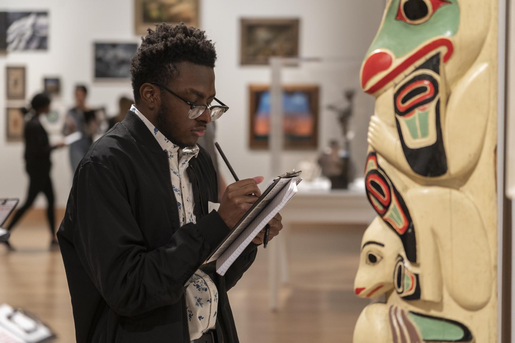 A young Black man wearing glasses writes on a clipboard while standing in front of a totem pole in a gallery