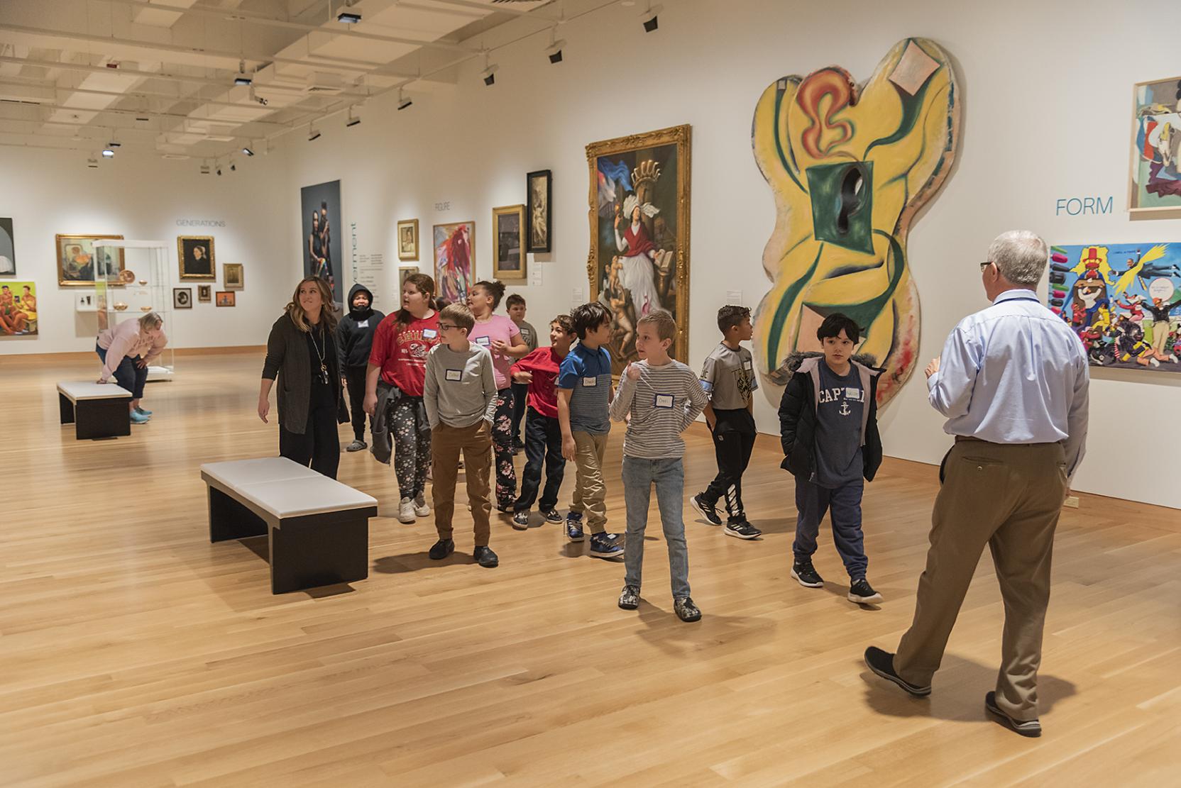 An older white man walks backwards while leading a group of elementary children through an art museum