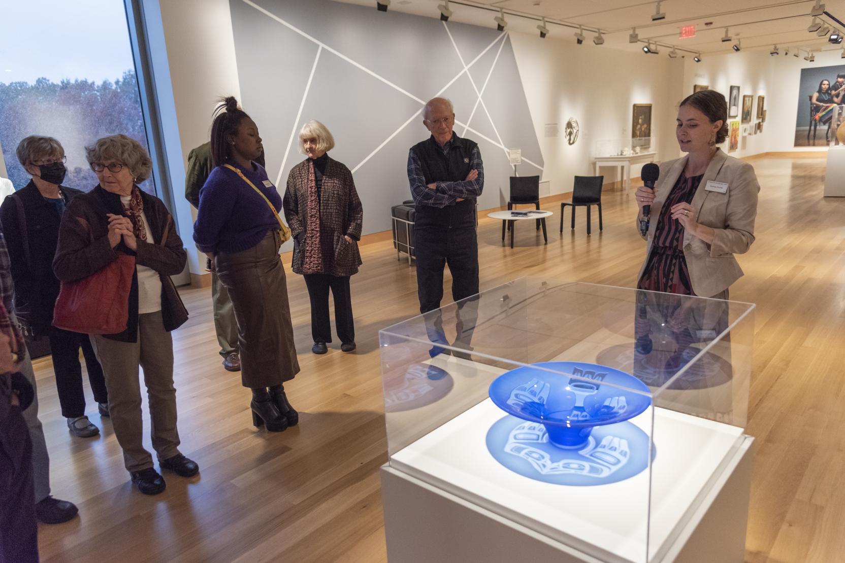 A woman with a microphone stands in an art gallery next to a blue bowl encased in glass; adults stand in front of her