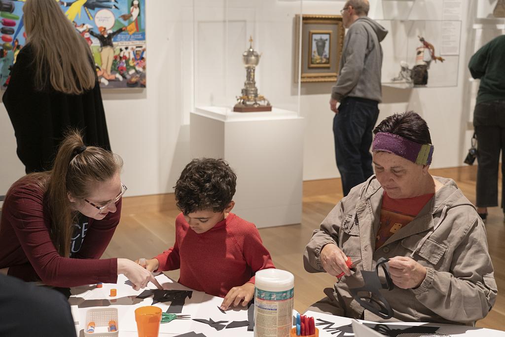 A woman, young boy, and older woman sit together in an art gallery at a table covered in craft supplies