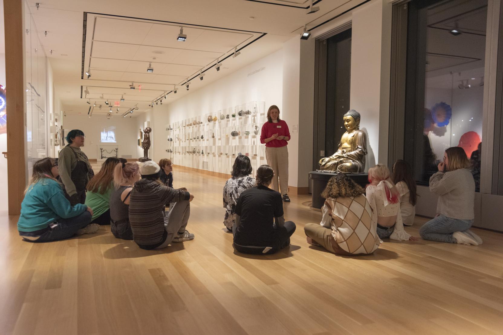 A young woman in stands next to a golden Buddha statue in a gallery space; a group of people sit in front of her