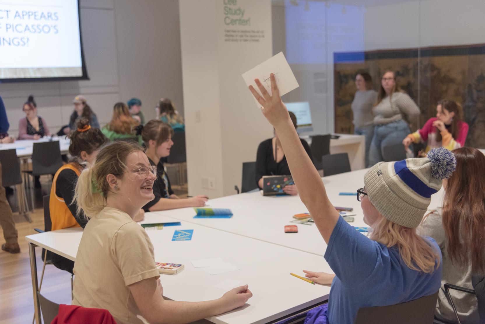 Students sit around a long table; one student holds a square piece of paper above her head