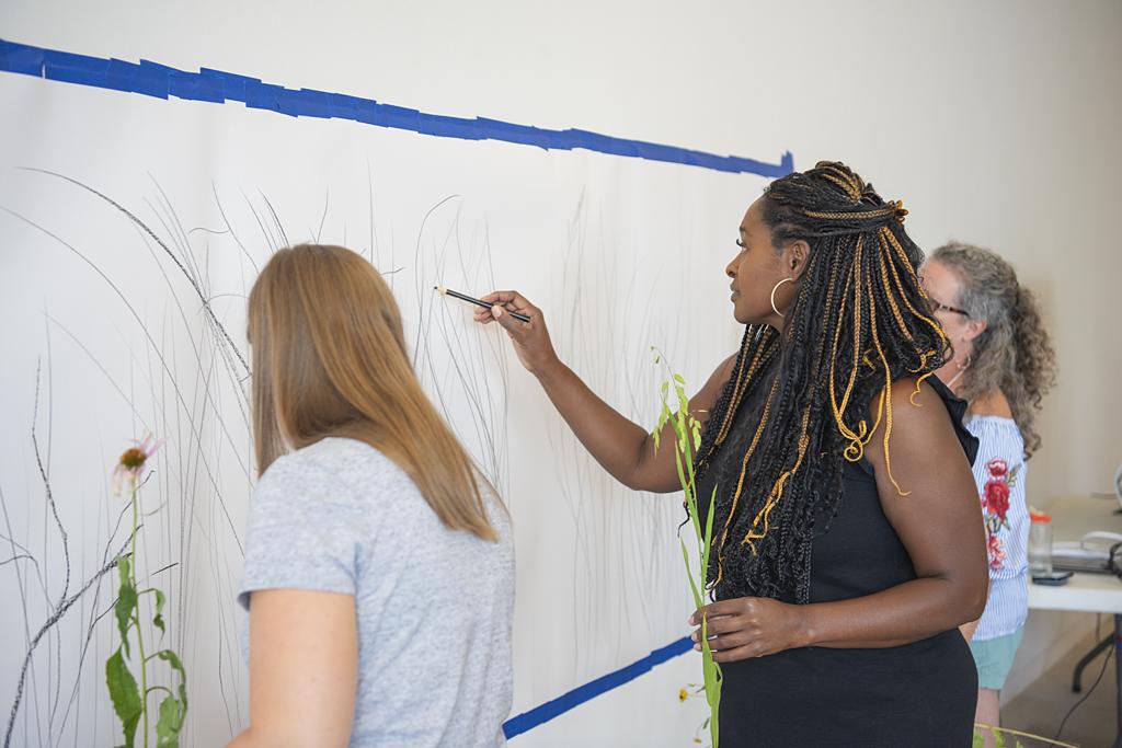 Two white women and one Black woman sketch flowers on a large paper taped to a wall
