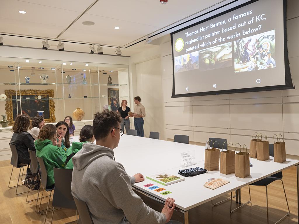 A group of young adults sit around a large white table facing a screen with a question and paintings on it