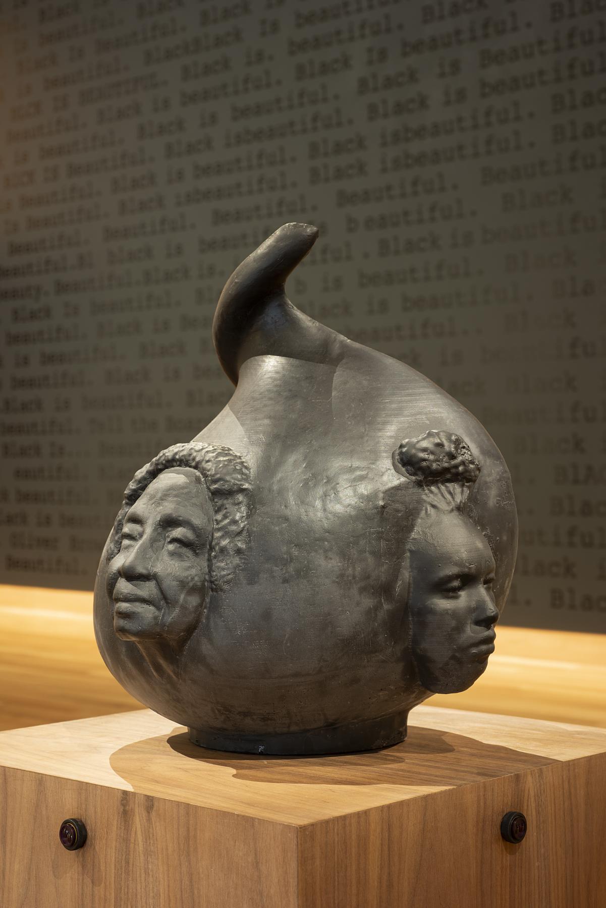 A black gourd-shaped sculpture with two female faces on it sits on a wooden platform in a gallery space