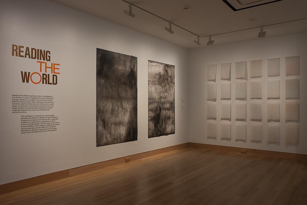 A corner of a gallery space; "reading the world” appears in red letters on the wall next to hanging artworks