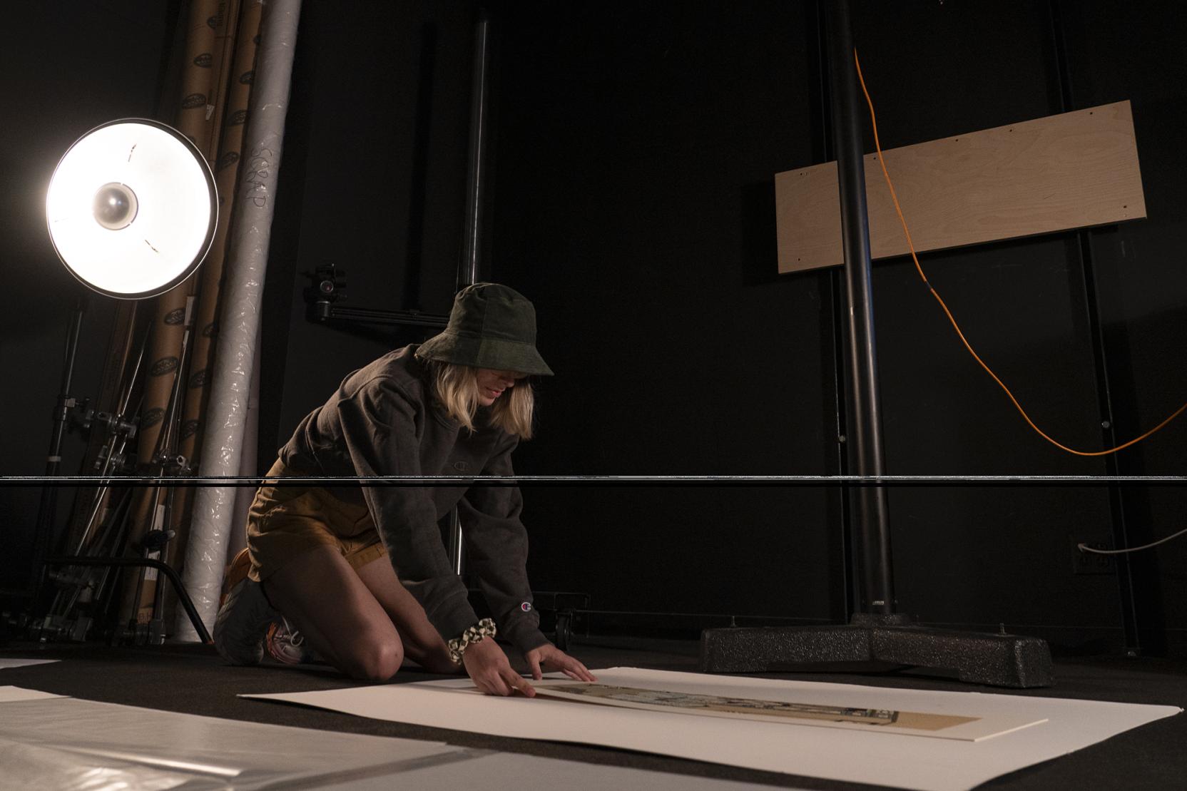 A student wearing a hat kneels on the ground in a dark room arranging papers with a bright lamp behind them