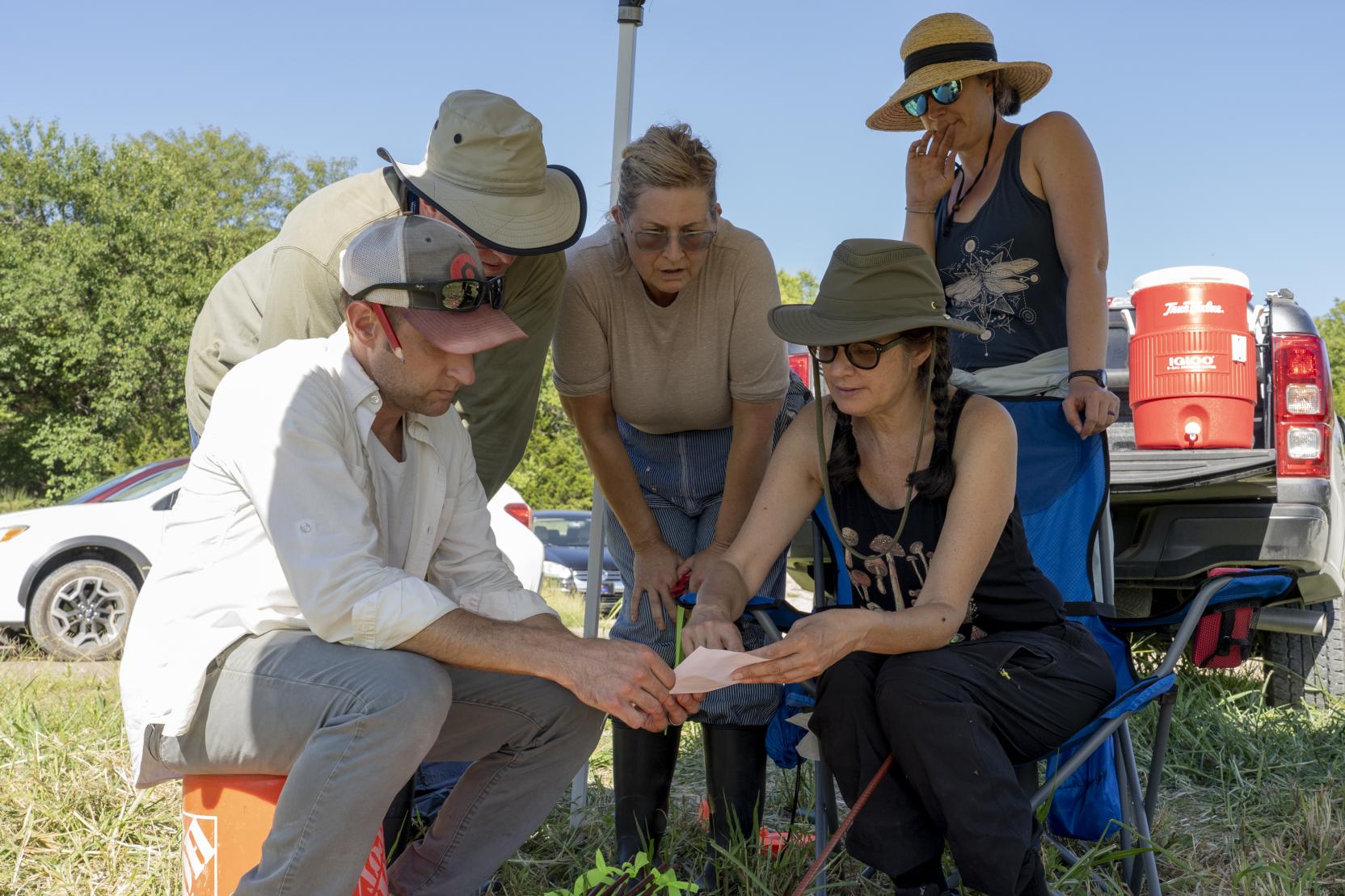 A white woman sits in a lawn chair and holds a piece of paper, four adults crouch around her looking toward the paper