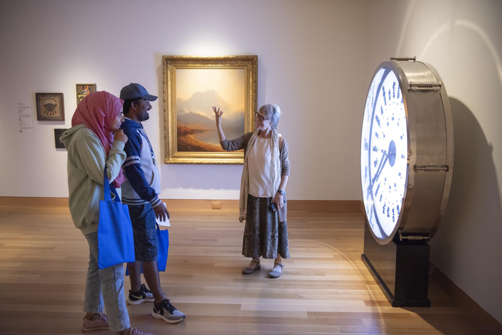 An older woman gestures in front of a large glowing clock in a museum gallery; two young adults stand in front of her
