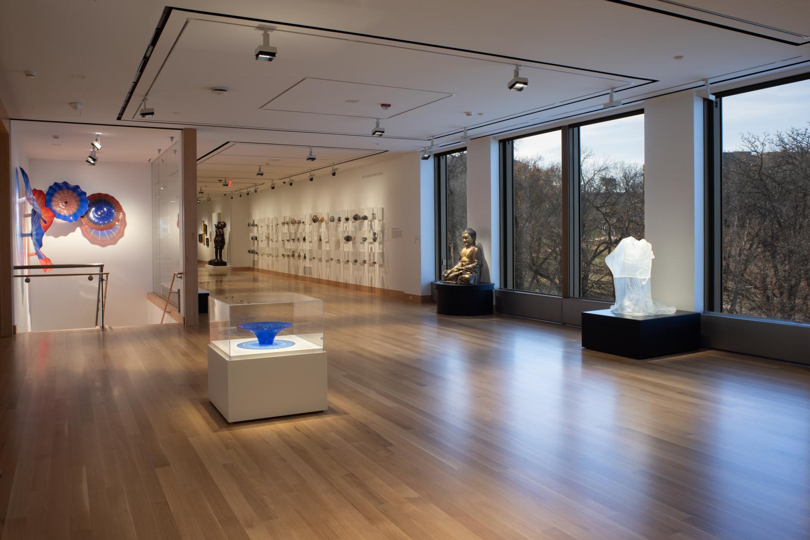 A dark gallery space with wooden floors, large windows on the right, and a variety of sculptures on display