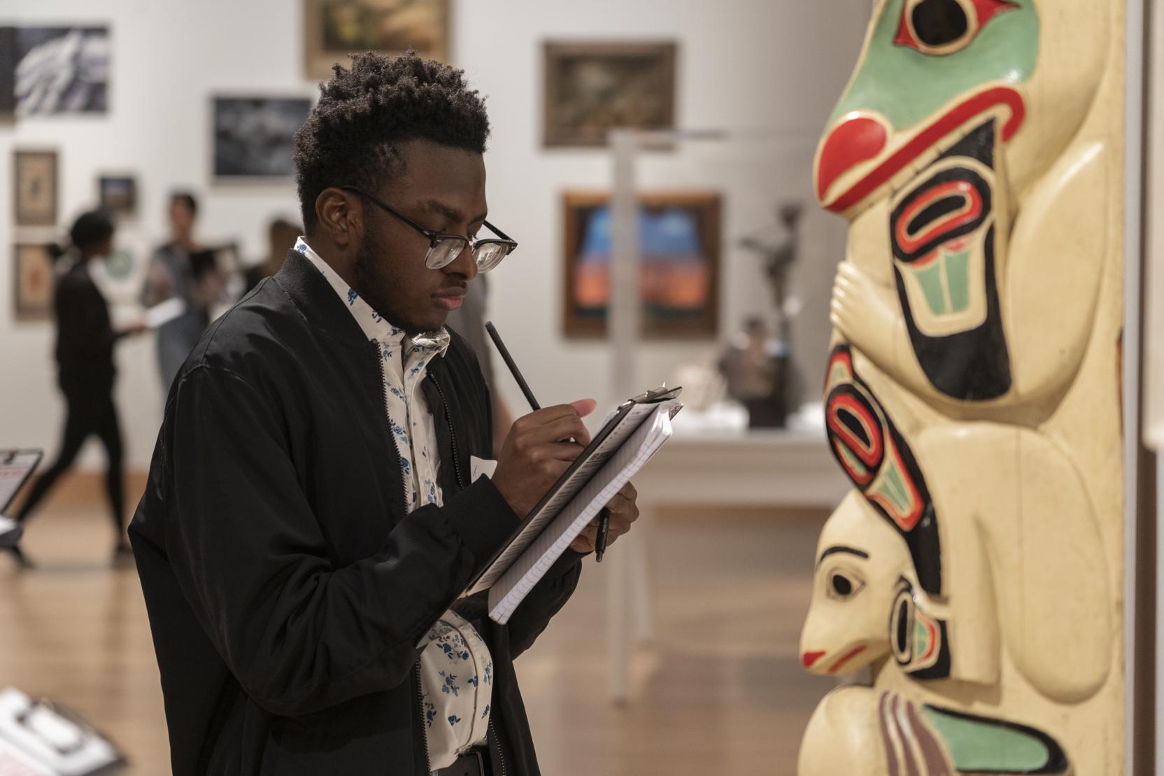 A young Black man wearing glasses writes on a clipboard while standing in front of a totem pole in a gallery