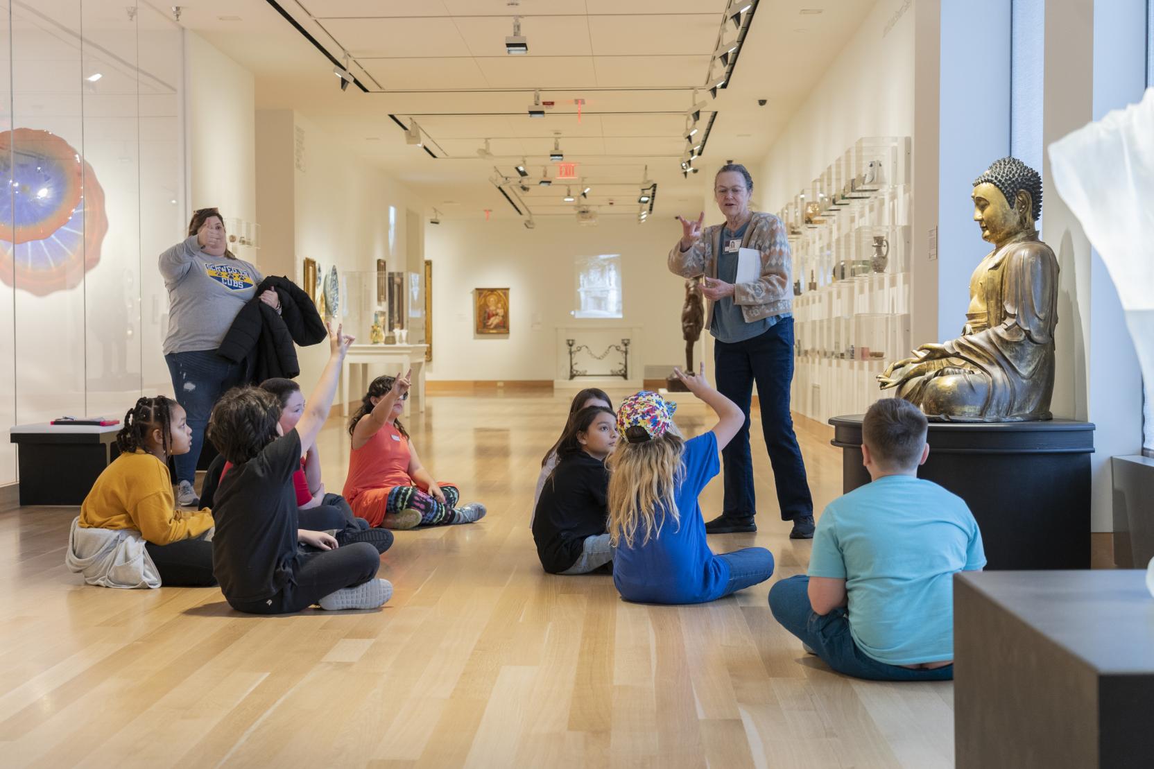 A group of children sit on the floor around a golden Buddha statue in an art gallery; two adults stand behind them