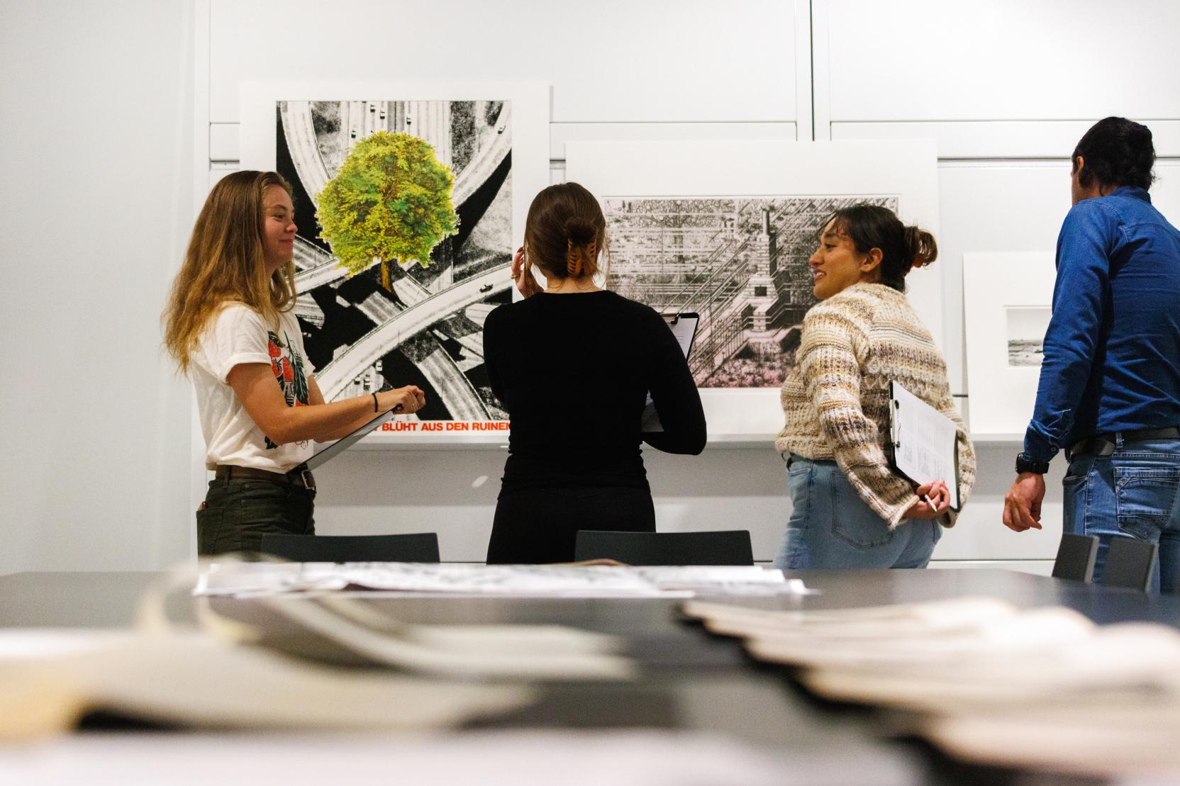 Four young adults in a bright classroom stand in front of two large art works propped against a white wall