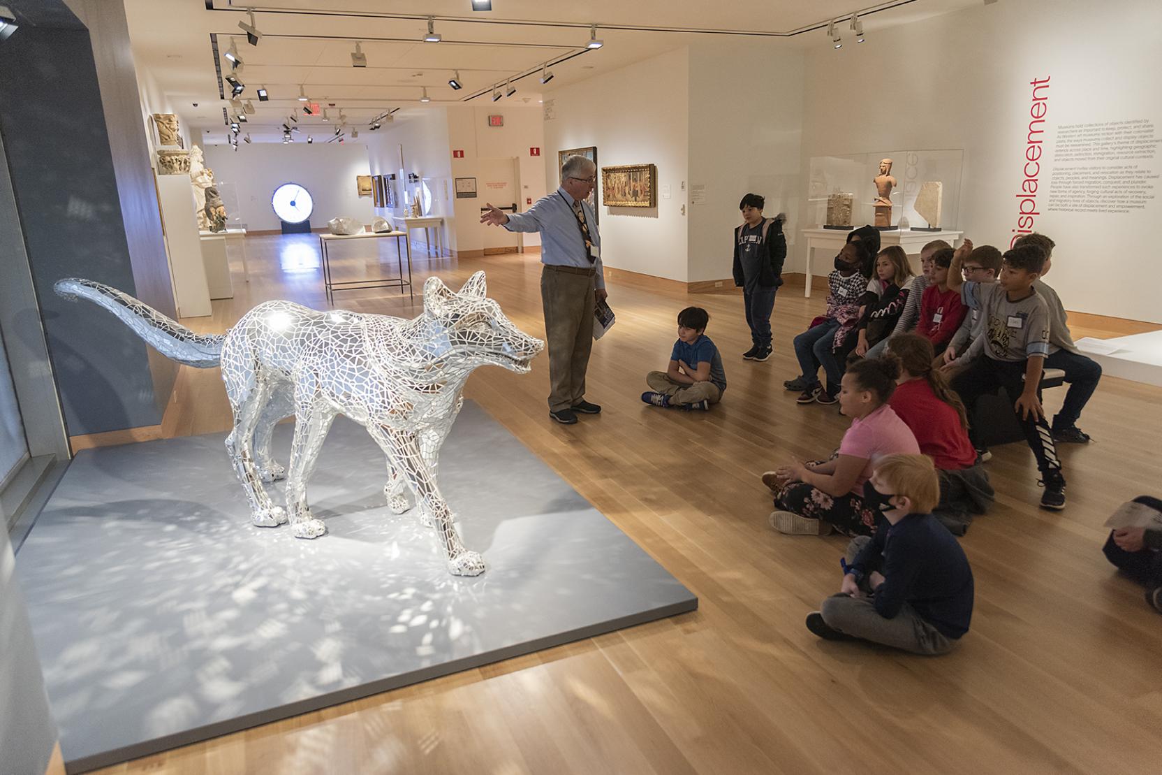 Children sit in an art gallery around a sculpture of a wolf covered in light reflective mirrors
