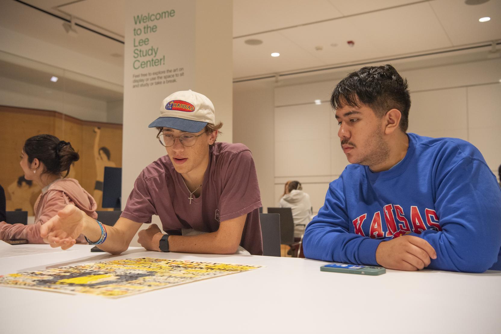 Two young men sit at a white table in a large classroom space looking over a yellow print