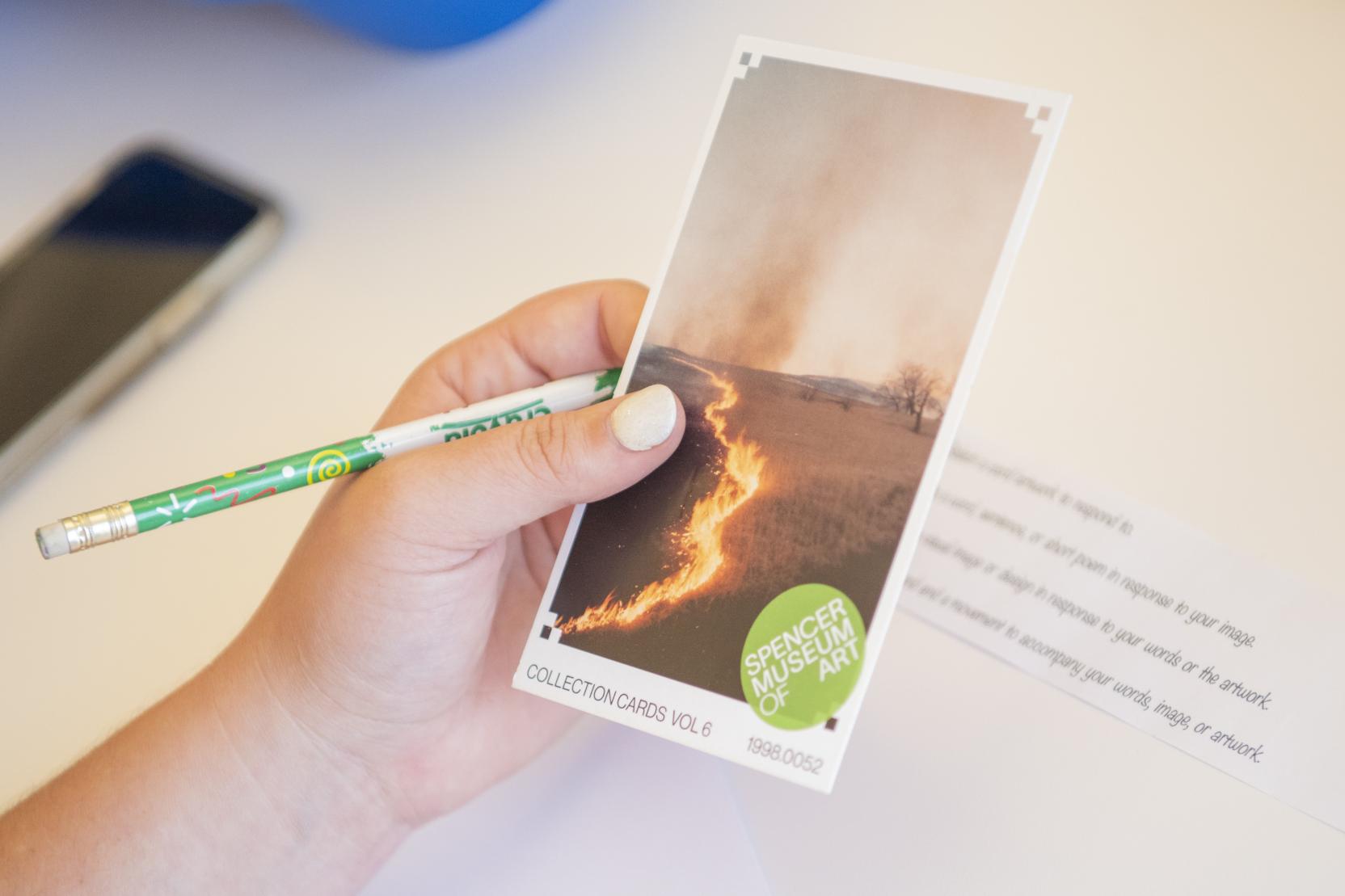 A white hand holds a pencil and a postcard with an image of a burning field on it