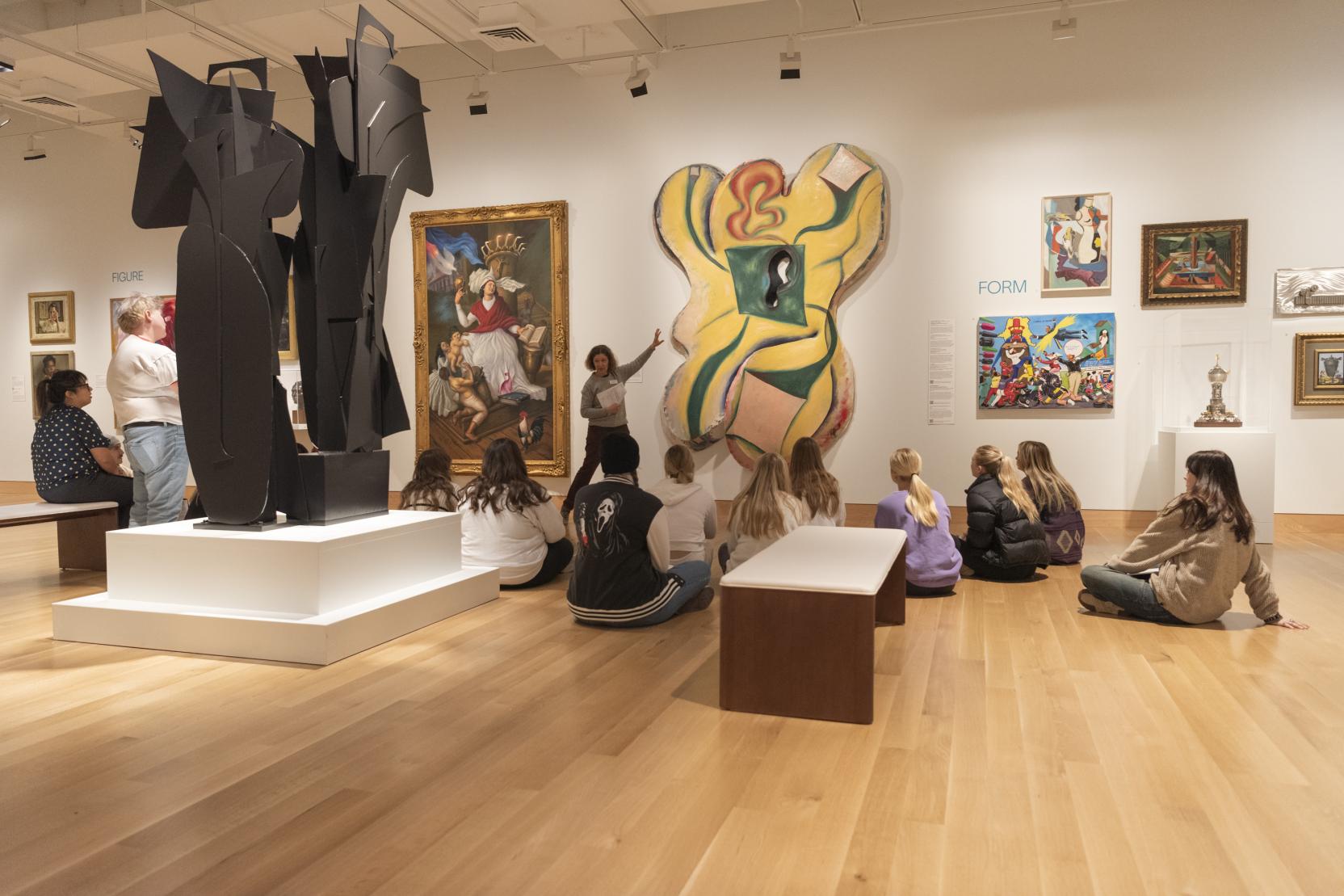 A group of children sit on the floor of an art gallery and look toward a woman gesturing to an artwork on the wall