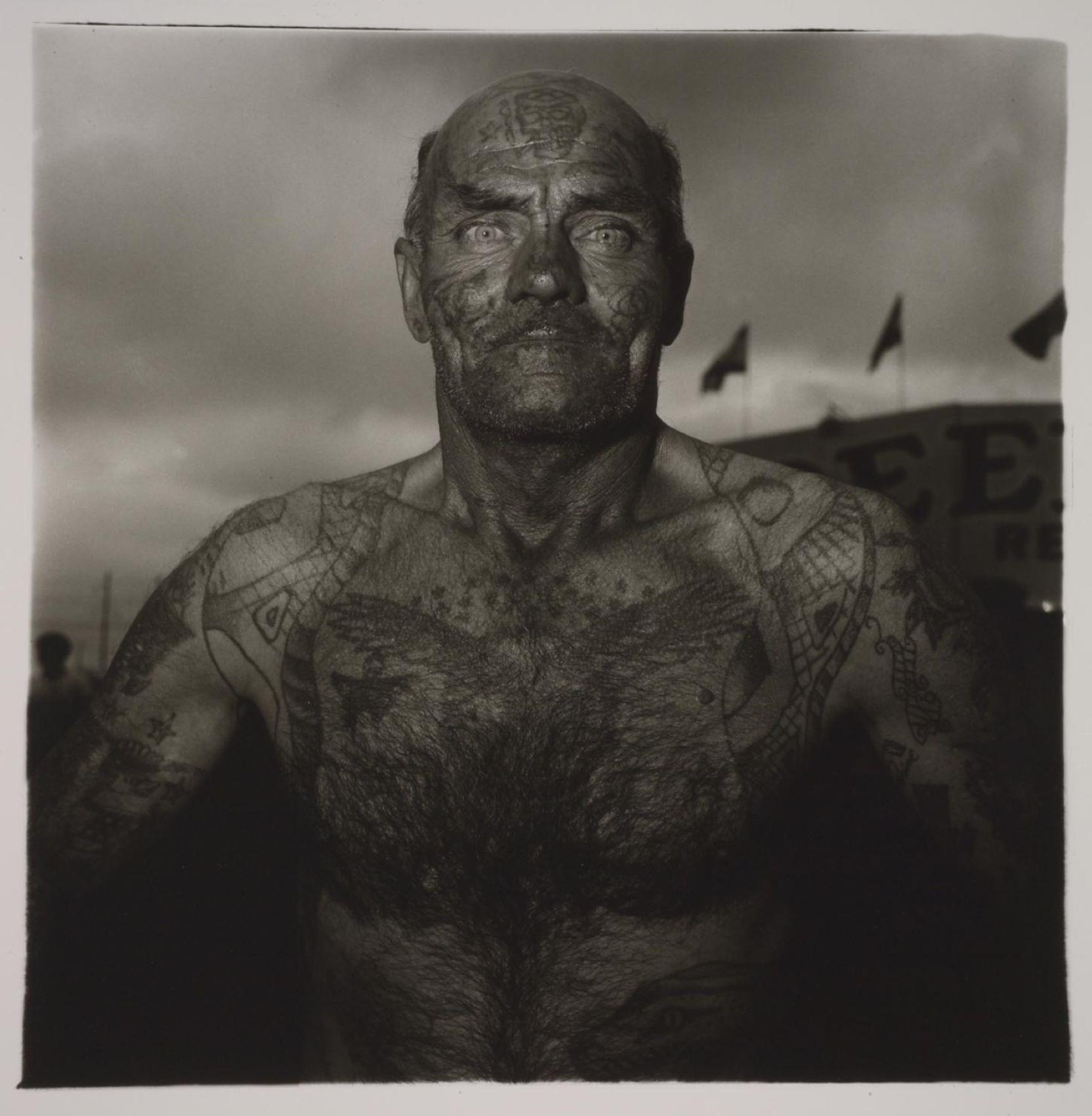 A black-and-white photo of a shirtless, balding white man with tattoos on his face and chest staring out toward the viewer
