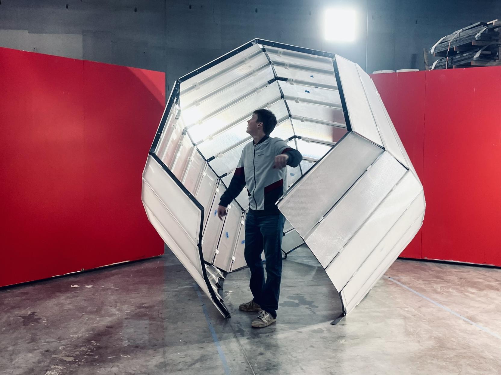 An adult stands inside a six-sided sculpture with an opening that rests on the ground of an industrial space